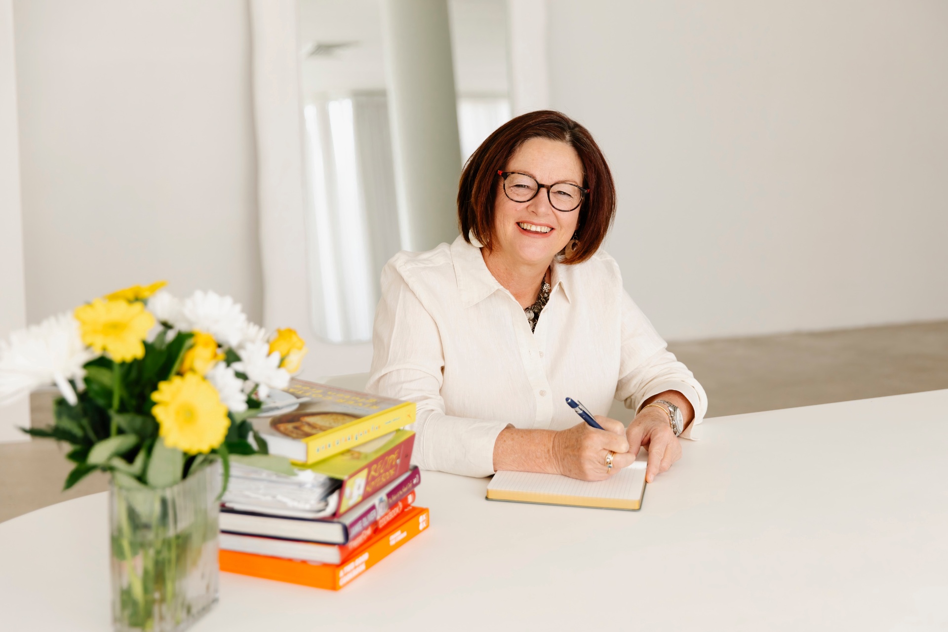 Sharon Hespe smiling while writing notes at a bright desk with cookbooks and flowers